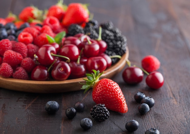 Fresh organic summer berries mix in round wooden tray on dark wooden table background Raspberries strawberries blueberries blackberries and cherries Top view