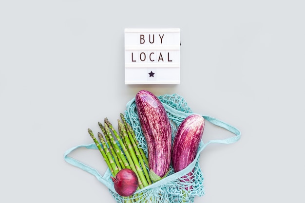 Fresh organic vegetables in eco cotton mesh shopping bag with text Buy Local on lightbox flat lay, top view with copy space on gray background. Sustainable lifestyle. Zero waste, plastic free concept.