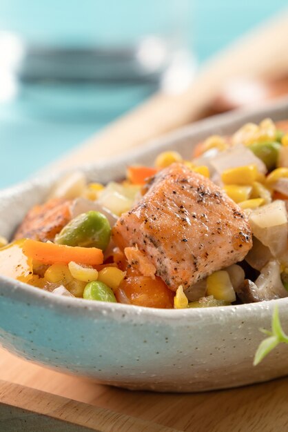 Fresh pan-fried salmon cube chops with stir-fried mixed vegetables including edamame beans, carrot, onion, mushroom and corn kernels on blue wooden table background.