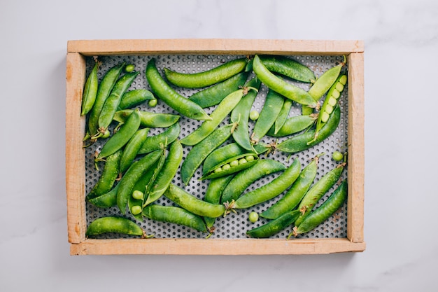 Fresh peas in a wooden tray on a marble background, flat lay, top view
