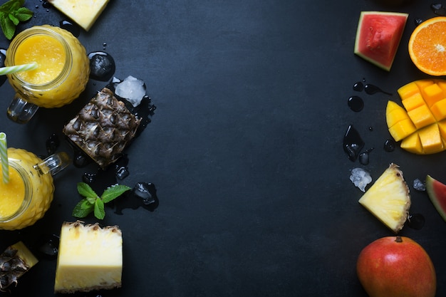 Fresh pineapple and mango smoothie in glasses with fruits on a black rustic background with copy space