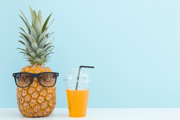 Fresh pineapple with sunglasses near a glass of juice and a cocktail straw on color background