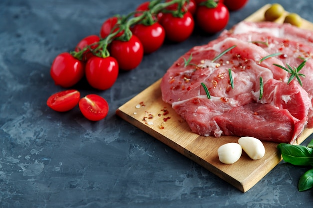 Foto braciole di maiale fresche in spezie su una tavola di legno, vicino a pomodorini su un ramo e spicchi d'aglio su uno sfondo scuro.