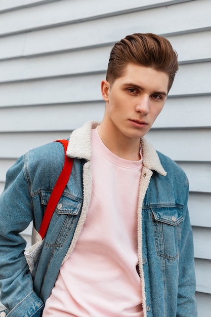 Fresh portrait European young man in vogue pink sweatshirt in fashionable blue denim jacket with fabric bag near vintage white building in city. Trendy nice guy fashion model outdoors. Street style.