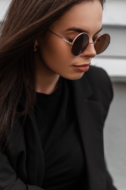 Фото Свежий портрет милой красивой молодой женщины в круглых модных солнцезащитных очках в черном стильном пиджаке с сексуальными губами на открытом воздухе в городе. привлекательная фотомодель девушки в повседневной элегантной деловой одежде.