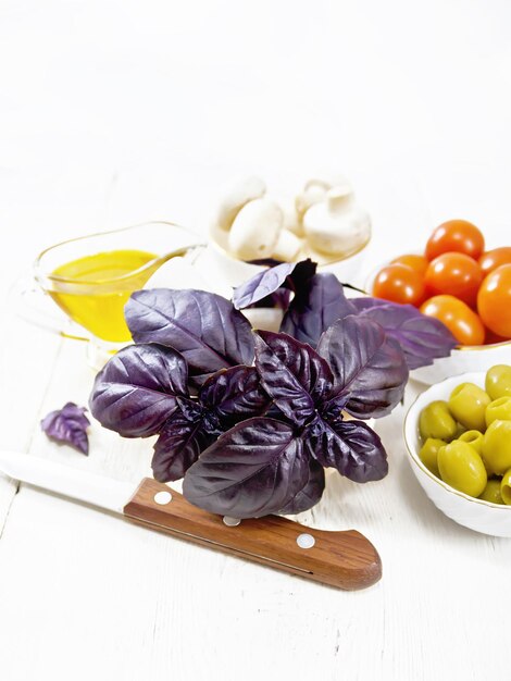 写真 モルタルオリーブトマトとシャンピニオンの新鮮な紫色のバジル3ボウルの植物油グレイビーボートと木の板の背景にナイフ