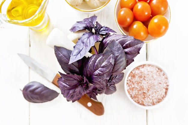 Fresh purple basil in a mortar pink salt tomatoes and champignons in bowls vegetable oil in gravy boat and a knife on wooden board background from above