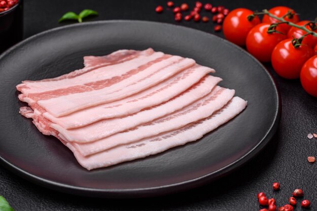 Fresh raw bacon cut into slices with salt spices and herbs on a dark textured concrete background