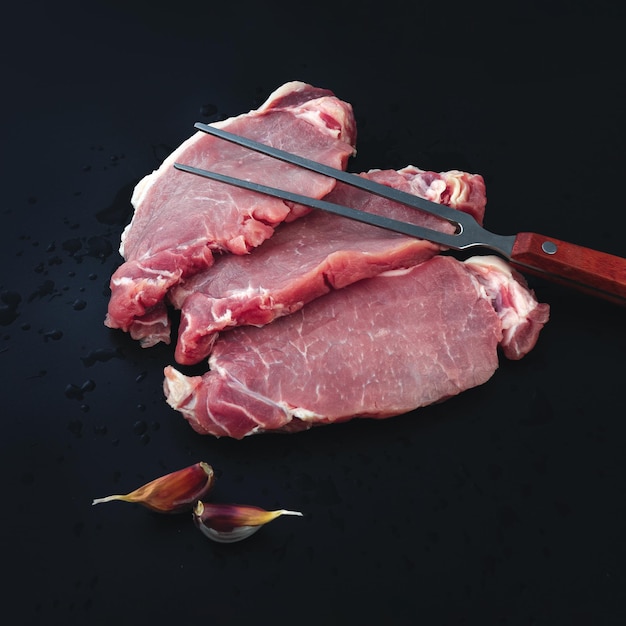 Fresh raw beef meat for grilling with seasoning and garlic and meat fork on dark background