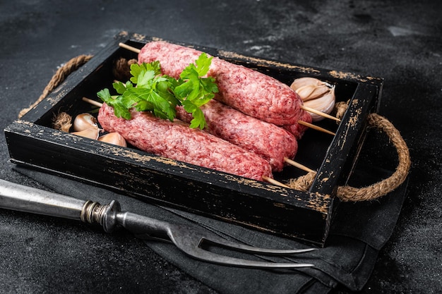 Fresh raw beef meat kebabs sausages on skewers in wooden tray Black background Top view