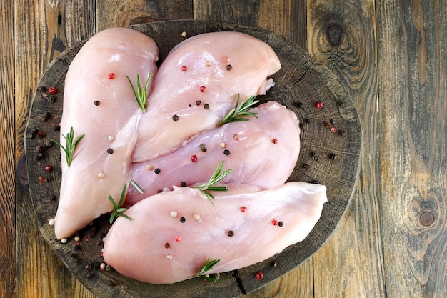 Fresh raw chicken breast fillets sprinkled with peppercorns and rosemary on round wooden board