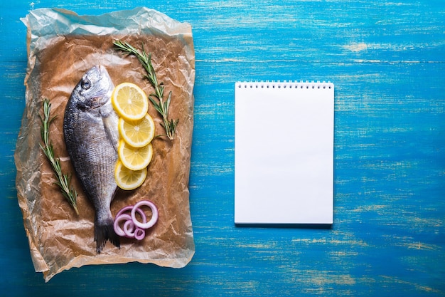 Fresh raw Dorado fish on cooking paper with spices and a notebook for recipe or menu. Top view on a blue background.