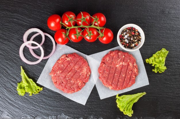 Fresh raw home-made minced beef steak burger with spices, tomatoes and salad on a black slate table, top view. Flat lay.