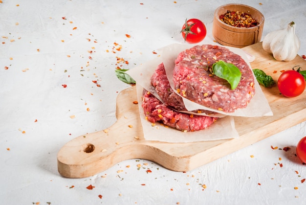 Fresh raw homemade minced beef steak burger with spices, tomatoes and basil, on a white stone concrete table, 