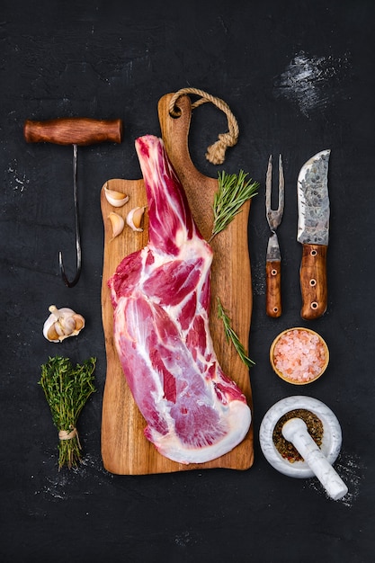 Fresh raw lamb leg bone in on wooden cutting board with herbs and seasoning