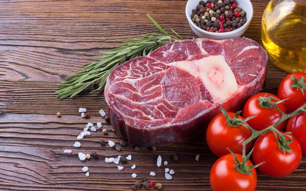 fresh raw meat beef steak with bone with spices, rosemary, tomatoes and olive oil on brown wooden background. cooking ingredients.