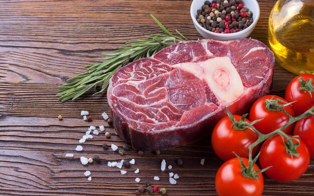 Fresh raw meat beef steak with bone with spices, rosemary, tomatoes and olive oil on brown wooden background. cooking ingredients.