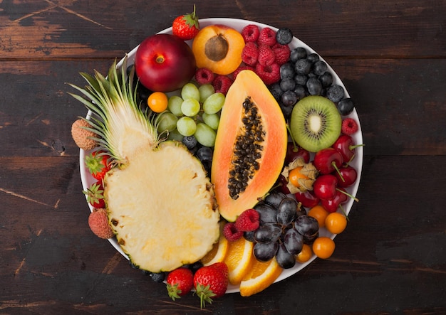 Фото Свежие сырые органические летние ягоды и экзотические фрукты в белой тарелке на темном деревянном фоне. ананас, папайя, виноград, нектарин, апельсин, абрикос, киви, клубника, личи, вишня. вид сверху