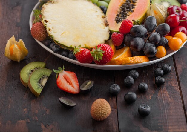 Fresh raw organic summer berries and exotic fruits in white plate on dark wooden background. Pineapple, papaya, grapes, nectarine, orange, apricot, kiwi, strawberry, lychees, cherry. Top view