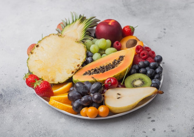 Fresh raw organic summer berries and exotic fruits in white plate on light background Pineapple papaya grapes nectarine orange apricot kiwi pear lychees cherry and physalis Top view