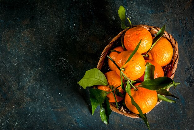 Fresh raw organic Tangerines with green leaves in little basket on dark concrete stone background, copy space