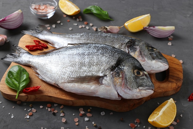 Fresh ready to cook raw fish dorado with ingredients and seasonings like basil, lemon, salt, pepper, red onion and garlic on wooden board on dark table. Closeup