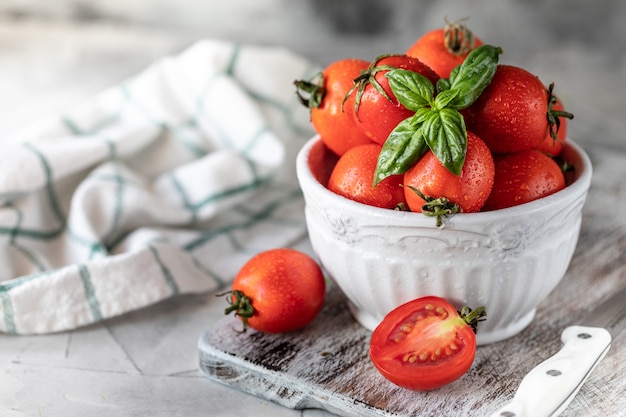 Fresh red cherry tomatoes and basil leaves in a plate on a white kitchen table.