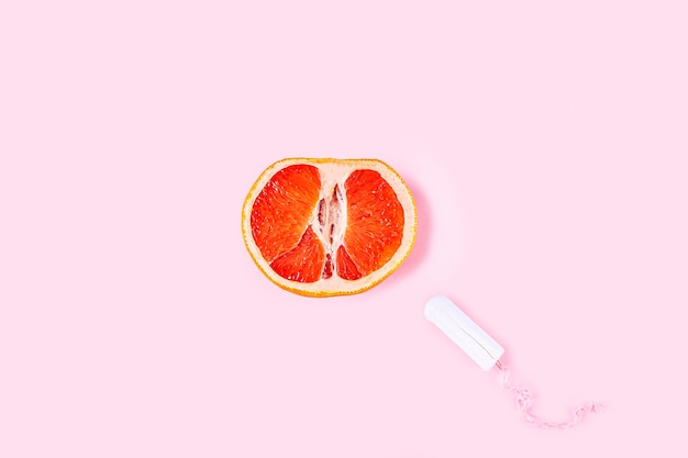 Fresh red cut grapefruit and tampon on pink background. Female health, Menstruation concept. Fruit as symbol of vagina. Close up.