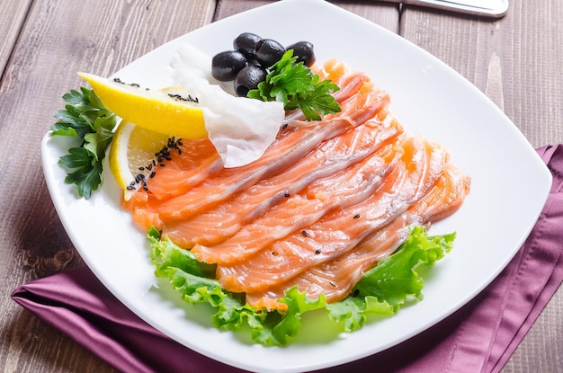 Fresh red fish cut into strips with lemon and herbs on a white\
plate on a dark wooden background