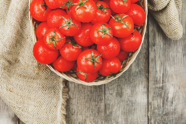 Pomodori rossi freschi in un cestino di vimini su una vecchia tabella di legno. pomodorini maturi e succosi con gocce di umidità, tavolo di legno grigio, intorno a un panno di tela. in stile rustico.