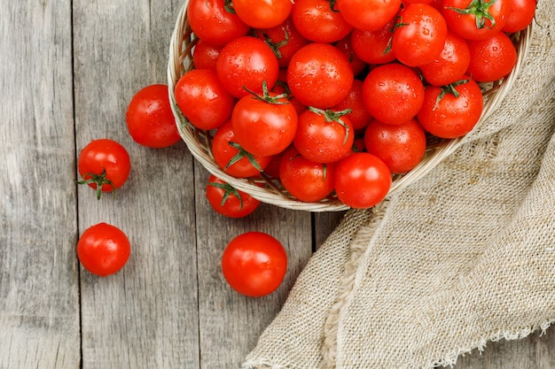 Fresh red tomatoes in a wicker basket on an old wooden table. Ripe and juicy cherry tomatoes with drops of moisture, gray wooden table, around a cloth of burlap. In a rustic style.