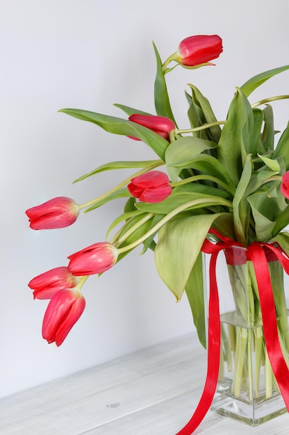 Fresh red tulips with green leaves and red ribbon in vase on white wooden background selective focus copy space greeting card for holidays of spring and loved ones