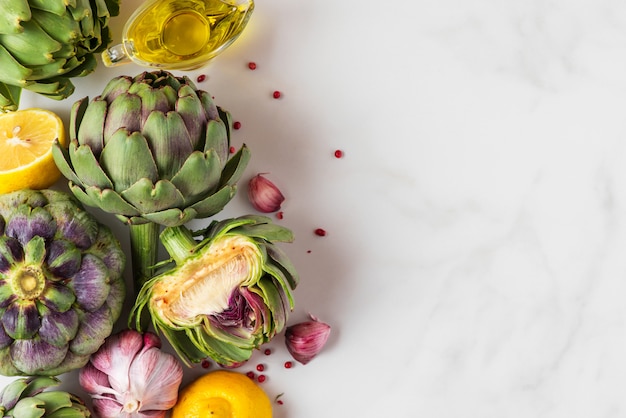 Carciofi freschi maturi, limone e aglio con olio d'oliva e pepe sul tavolo di marmo bianco.