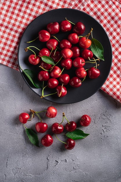 Fresh ripe cherry fruits with green leaves on black plate with red dish towel  summer vitamin berries on grey stone background, 