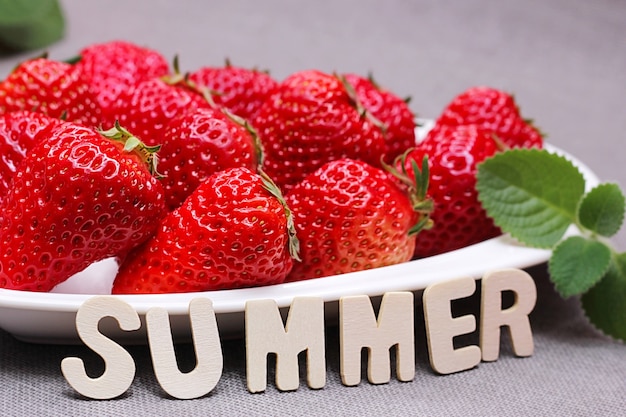 Fresh ripe delicious red strawberries and a green mint leaf on a white heart-shaped plate. the word summer is laid out in small wooden letters next to it