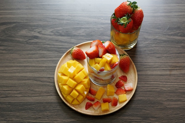 Fresh ripe mango and strawberry fruit in a cup with cookies soy yogurt on a wooden plate