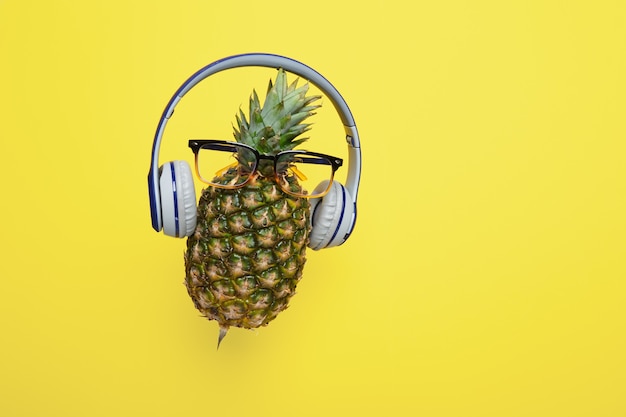 A fresh ripe pineapple in eyeglasses wearing wireless headphones on yellow background