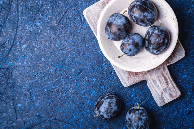 Fresh ripe plum fruits in white wooden bowl and old cutting board on blue abstract background, top view copy space