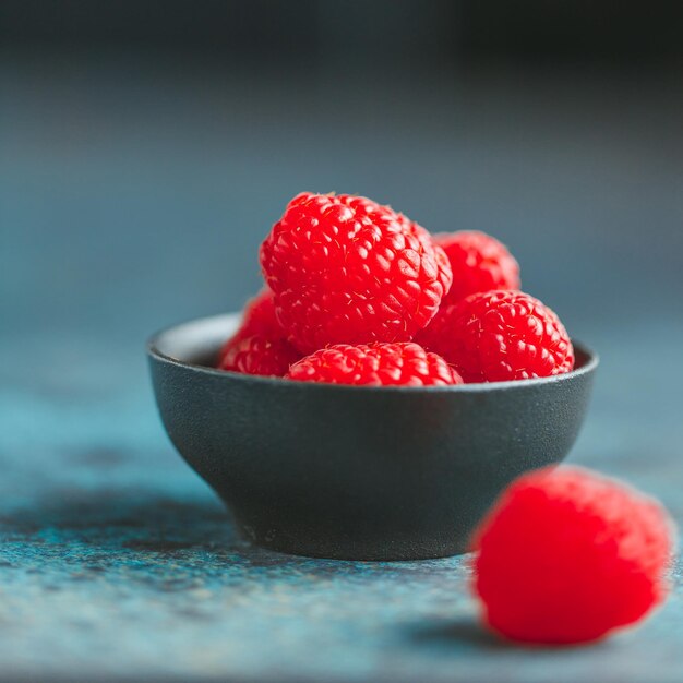 Fresh ripe raspberries in a small black bowl on a textured blue table Macro photography