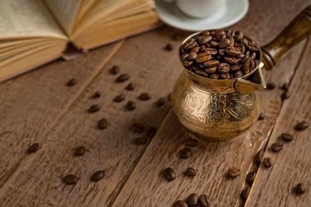 Fresh roasted coffe beans in  cezve (traditional turkish coffee pot) opened book and cup on wooden table.