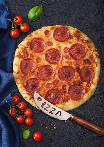 Fresh round baked Pepperoni italian pizza with knife with tomatoes and basil on black kitchen table background