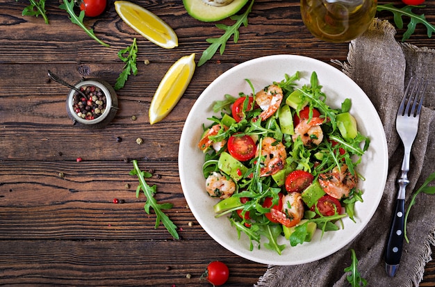 Fresh salad bowl with shrimp, tomato, avocado and arugula on wooden table close up. Healthy food. Clean eating. Top view. Flat lay.