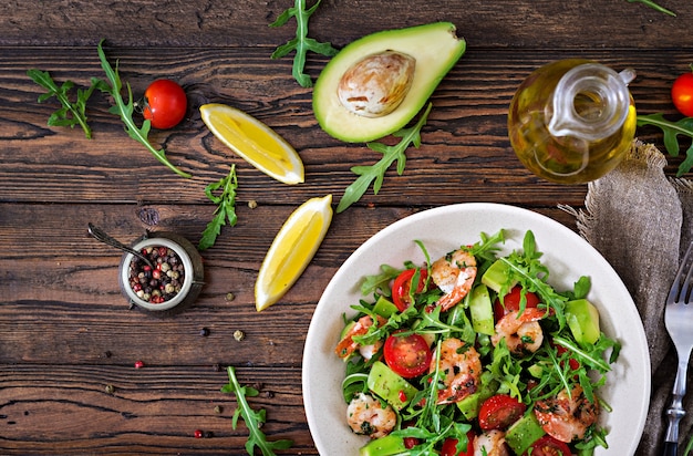 Fresh salad bowl with shrimp, tomato, avocado and arugula on wooden table close up. Healthy food. Clean eating. Top view. Flat lay.