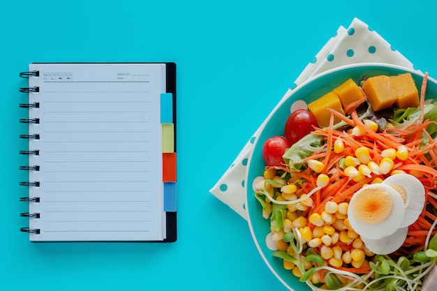 Fresh salad vegetable with boiled chicken egg and blank spiral notebook on blue background