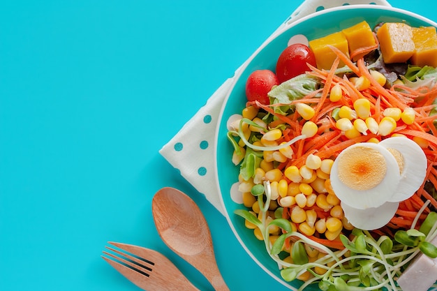 Fresh salad vegetable with boiled chicken egg, wooden spoon and fork on blue background