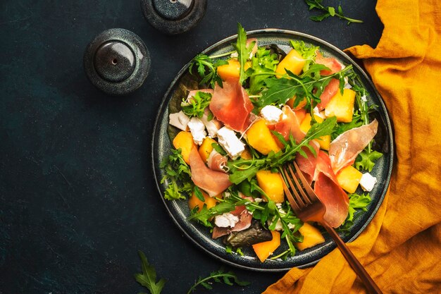 Fresh salad with cantaloupe melon prosciutto soft cheese and arugula on blue background top view copy space