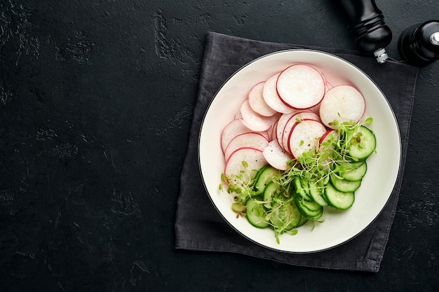 Fresh salad with red radish, cucumber, vegetables, microgreen radishes in white plate on grey stone background. View from above. Concept vegan and healthy eating.