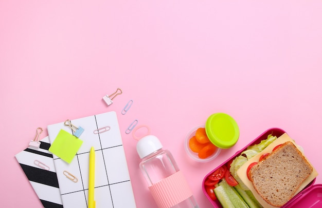 Panino, patata e cetriolo freschi in scatola di pranzo rosa con la bottiglia di acqua nel luogo di lavoro dell'ufficio primo piano dello spuntino sano in recipiente di plastica concetto sano dell'alimento vista superiore, disposizione piana, fondo rosa.