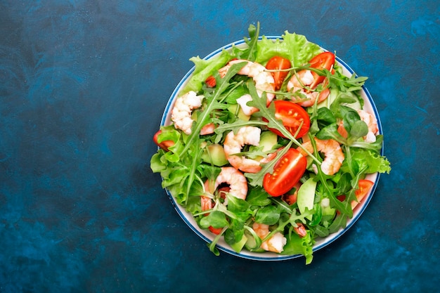 Fresh shrimp salad with tomatoes lettuce arugula avocado cucumber and lemon dressing on blue background Healthy eating clean food concept Top view