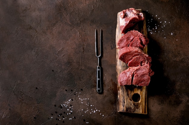 Fresh sliced raw beef tenderloin meat for steaks on wooden board with metal meat fork, salt, pepper over dark brown texture surface. Food cooking background concept. Top view, flat lay, Copy space